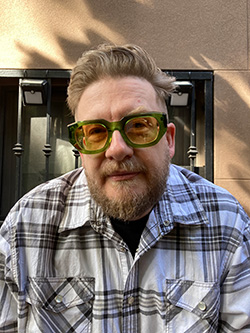 Justin Neely shown in medium closeup wearing tinted glasses and a plaid shirt