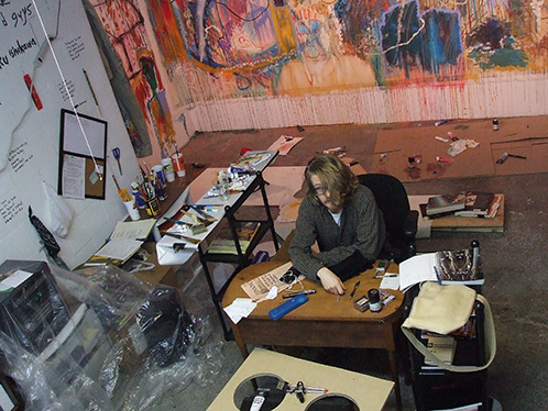 justin neely sits at a desk in an art studio with paintings on wall behind him